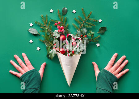 Creative Christmas flat lay on green paper. Hands and red Xmas decorations in veneer cone - fir and holly twigs, baubles, candy canes, wooden stars an Stock Photo