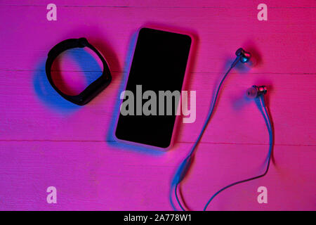 Top view of set of gadgets in purple neon light and pink background. Smartphone and smartwatch, headphones. Copyspace for your advertising. Tech, modern, gadgets. Stock Photo