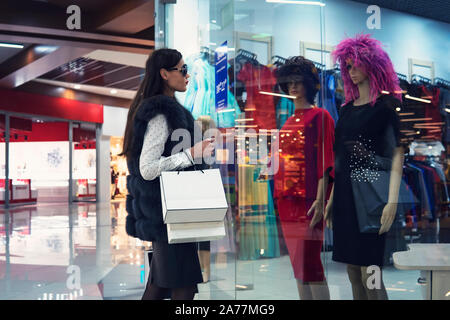 Young woman in shopping centre standing near the shop window and looking at two mannequins in dresses and wigs. Trendy attractive girl is doing shoppi Stock Photo