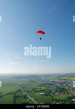 This skydiver is flying high under his canopy in the blue sky. He has save opened his parachute and soaring down to the dedicated landing area. Stock Photo