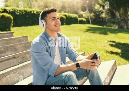 Outdoors Leisure. Stylish guy in headphones wearing watch sitting on stairs in park with smartphone listening to music smiling happy Stock Photo