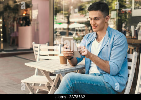 Outdoors Leisure. Stylish guy sitting at cafe on the street with cup of coffee using digital tablet smiling joyful Stock Photo