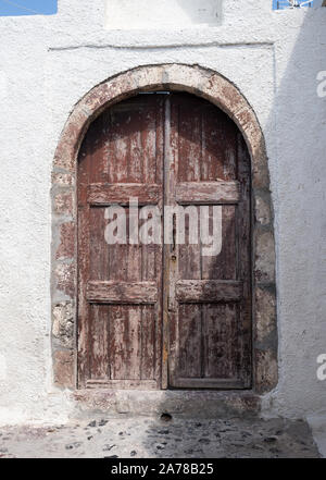Old brown curved top arch peeling panel door in white wash concrete wall Fira Santorini Stock Photo