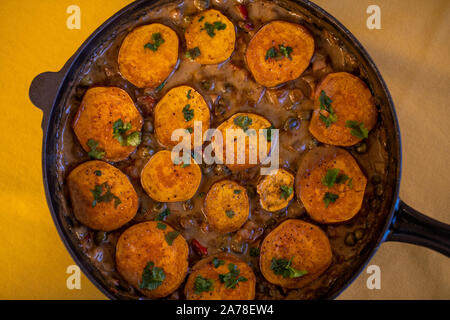 Baked sweet potatoe and pea curry.  Baked sweet potatoe with cooked vegetables, served in an iron cast pan. Plant based vegan food. Stock Photo