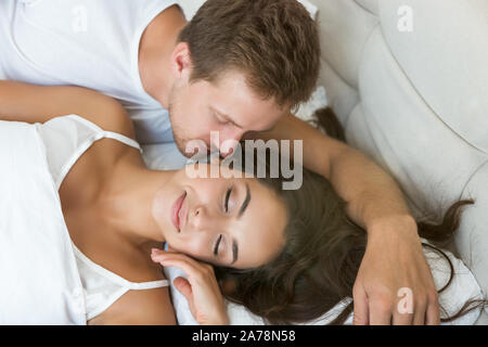 young couple handsome husband kissing his beautiful sleeping wife in bed in bright bedroomfeeling happy family morning idyll Stock Photo