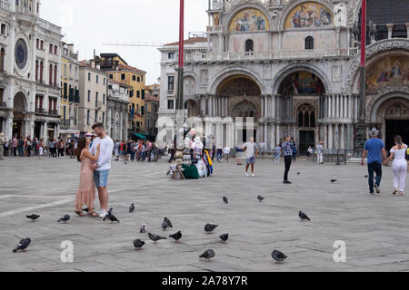 Young romantic couple standing, holding each other, in St Marks Square surrounded by pigeons and a backdrop of tourists and famous buildings. Stock Photo