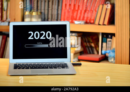 on the table laptop with text - 2020 loading - on screen, blurred shelf with books in the background - image Stock Photo