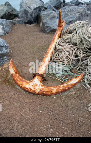Large rusty anchor on sandy beach with rope coiled up beside it, rocks behind. Stock Photo