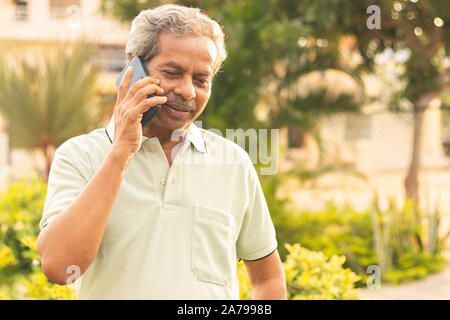 Healthy senior Asian on phone outdoor green park - Elder man busy in mobile - Happy male parent talking on phone. Stock Photo