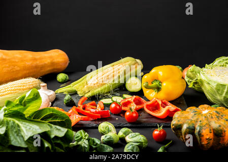 Chopped vegetables on a black board and different fresh vegetables on a black table. Stock Photo