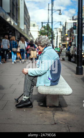A full length portrait of an old woman sitting on a street bench, Oxford Circus, London, England, UK. Stock Photo