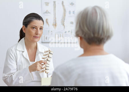 Aging and back pain concept: doctor of orthopedics showing her senior patient a slipped disk on a backbone model. Stock Photo