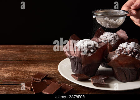 Still life on old wooden board table with three cocoa muffins with sugar and pieces of chocolate. Female hand holding strainer on black background. Sp Stock Photo