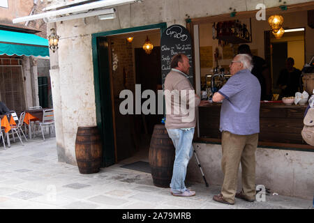 Two men, one older, standing outside a traditional style Venetian bar/cafe casually chatting together. Another cafe with chairs in the background. Stock Photo