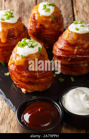 Baked potatoes wrapped in bacon topped with cheese and sour cream close-up on the table. vertical Stock Photo