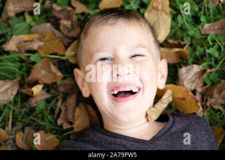 Portrait of a happy young pre-school boy lying on the grass and dry leaves and laughing in public park in autumn. Caucasian boy having fun in autumn. Stock Photo