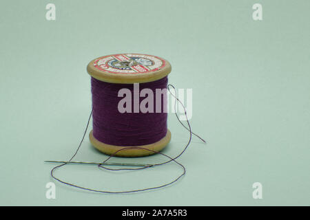Old wooden cotton reel wound with purple thread on white background with needle in foreground, label on top of reel Stock Photo
