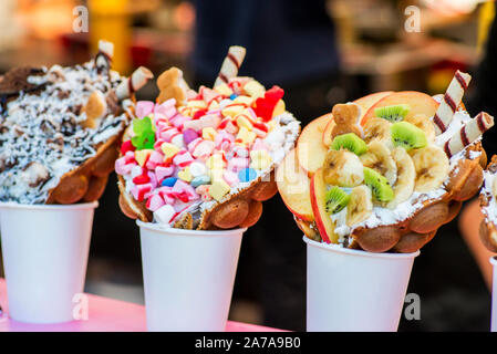 Bubble waffles with raspberries, chocolate and marshmallow in white paper cups are sold on Open kitchen food festival event. Street food in food stall Stock Photo