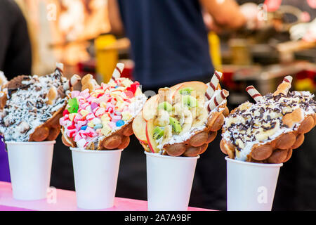 Bubble waffles with raspberries, chocolate and marshmallow in white paper cups are sold on Open kitchen food festival event. Street food in food stall Stock Photo