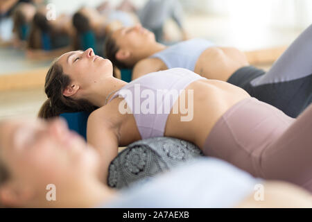 Restorative yoga with a bolster. Group of three young sporty attractive women in yoga studio, lying on bolster cushion, stretching and relaxing during Stock Photo