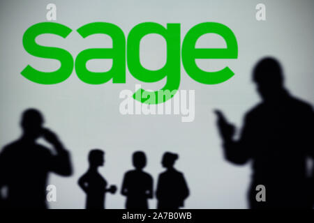 The Sage Group logo is seen on an LED screen in the background while a silhouetted person uses a smartphone (Editorial use only) Stock Photo
