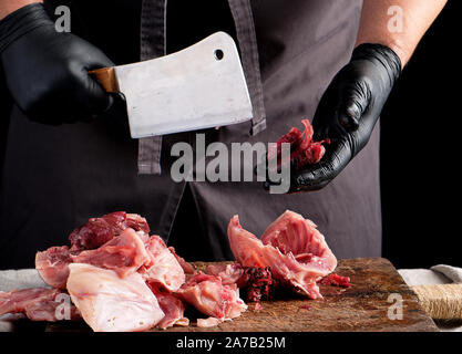 chef in black latex chunks cuts into pieces raw rabbit meat on a brown wooden board, cooking on a dark background Stock Photo