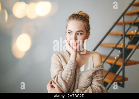 A young beautiful thoughtful girl with blonde hair in an evening silk dress and a cozy cardigan is dreaming about something at home against the backgr Stock Photo