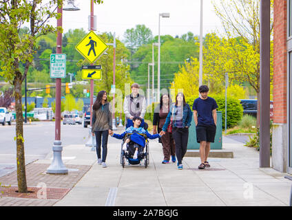 Disabled biracial boy in wheelchair walking along city street with father, siblings and caregiver, holding hands Stock Photo