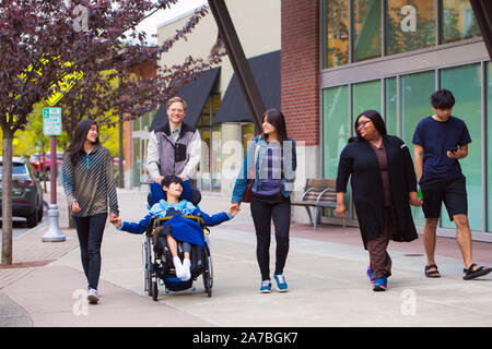 Disabled biracial boy in wheelchair walking along city street with father, siblings and caregiver, holding hands Stock Photo