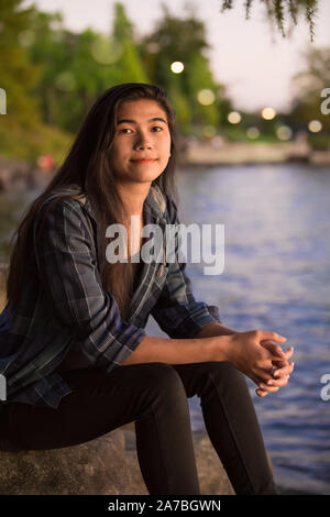 Biracial Asian Caucasian young teen girl sitting by edge of lake on rocks at sunset Stock Photo