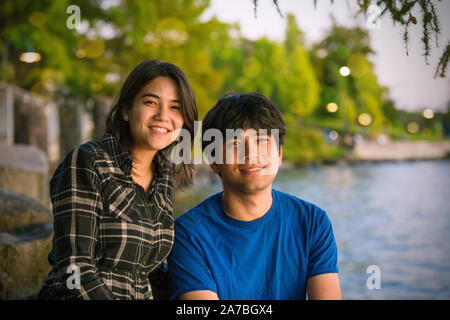 Young biracial Asian Caucasian adults sitting together outdoors by lake near dusk or sunset, smiling together Stock Photo