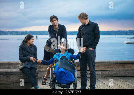 Mixed race family with disabled little boy in wheelchair outdoors on wooden pier by lake on cloudy evening at sunset in early autumn Stock Photo
