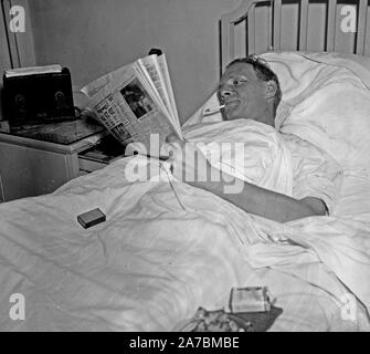 Man in hospital bed smoking a cigarette while reading a newspaper ca. 1936 Stock Photo