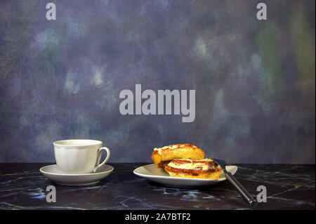 A white ceramic cup with black coffee and a plate with two cinnamon rolls in icing and a fork on the marble table. Close-up. Stock Photo