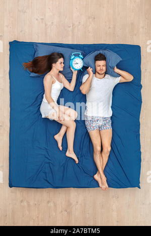 Young couple man covering ears with pillow trying to sleep irritated while woman holding alarm clock waking him up concerned lying on bed top view Stock Photo