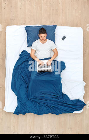 Young man lying on bed under blanket bedtime top view working on laptop online typing concentrated Stock Photo