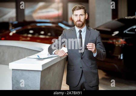 Portrait of a confident sales manager in the suit standing with car key in the showroom with electric cars on the background Stock Photo