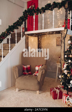 View on comfy brown armchair with two checked pillows standing in beautifully decorated Christmas living room with fir tree, presents and stairs with Stock Photo