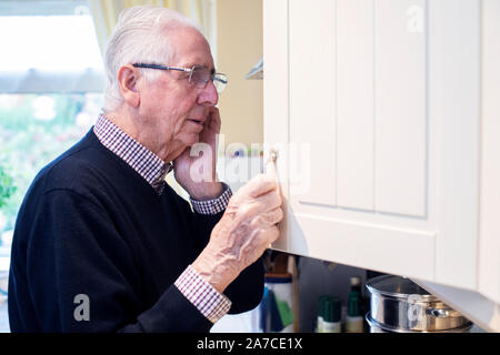 Forgetful Senior Man With Dementia Looking In Cupboard At Home Stock Photo