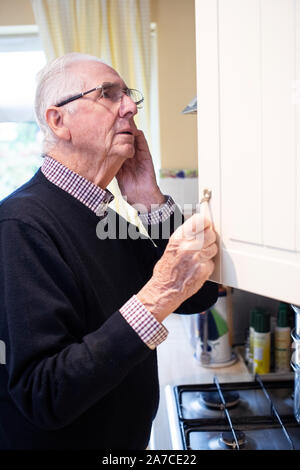 Forgetful Senior Man With Dementia Looking In Cupboard At Home Stock Photo