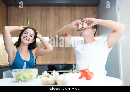 young funny couple cooking breakfast in the kitchen man closes his eyes with mushrooms while his beloved wife holds green onion like ears laughing Stock Photo