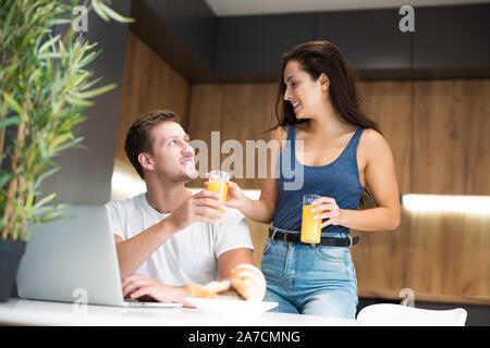 young lovely couple having breakfast woman passes her husband glass with orange juice while he works in laptop in kitchen great start of day Stock Photo