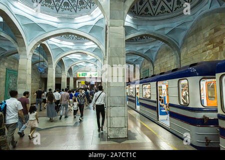 A number of commuters walking down the platform towards the exit of Alisher Navoi Metro Tashkent as a train sits at the station with its doors open. Stock Photo