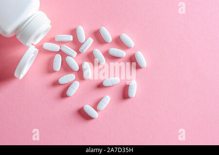 A white plastic container with white tablets lies on a multi-colored background next to white tablets Stock Photo