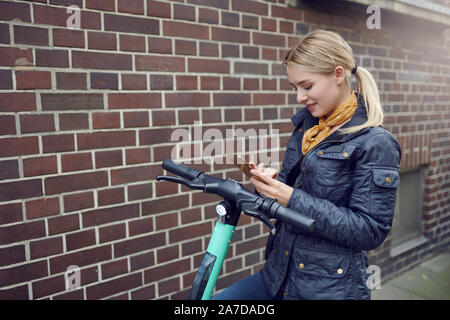 Trendy young blond woman in leather jacket standing at her e-scooter booking a ride, ecological transportation concept with copy space Stock Photo