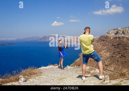 Young  man photographs a beautiful woman on the background of the sea and the island of Santorini Stock Photo