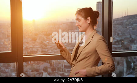 Young successful businesswoman in office suit is browsing mobile phone, side view. She is standing near the window with panoramic city view in her office in evening, sunlight Stock Photo