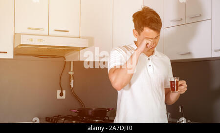 Portrait of tired and sleepy young man is drinking a tea in the morning in kitchen at home. He yawns and rubs his eyes, trying to wake up. Early morning on a weekday. Stock Photo