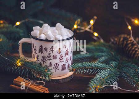 Fir tree colored ceramic mug with coffee or hot chocolate with marshmallows on bright wooden background with glittering garland Stock Photo