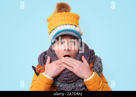 Handsome caucasian shocked funny man in several hats and scarfs being surprised on blue background. Winter fashion. Stock Photo